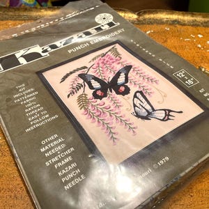 May include: A vintage Kazari punch embroidery kit featuring a framed butterfly design with pink wisteria. The kit includes patterned fabric, rayon thread, and instructions. The finished piece measures 12 x 16 inches.