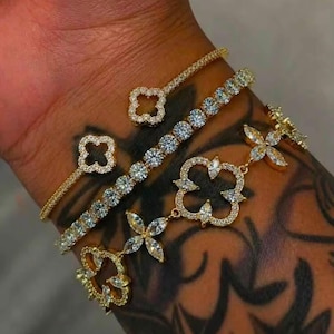 Könnte beinhalten: Drei goldfarbene Armbänder mit klaren Steinen. Eines ist ein offener Armreif mit kleeblattförmigen Enden, eines ist ein Tennisarmband mit runden Steinen und eines ist ein Gliederarmband mit kleeblattförmigen Motiven aus Markisen- und Rundsteinen.