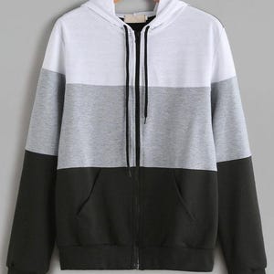 Op de afbeelding: Een colorblock hoodie met een wit bovenstuk, een grijs middenstuk en een zwart onderstuk. De hoodie heeft een volledige ritssluiting, een capuchon en een kangoeroezak. Het kledingstuk hangt aan een houten hanger.