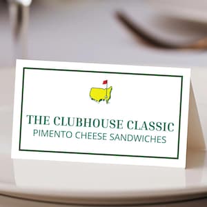 May include: A white card with a green border and text that reads "THE CLUBHOUSE CLASSIC PIMENTO CHEESE SANDWICHES." The card features a yellow outline of a golf course with a red flag. The card is on a white plate.