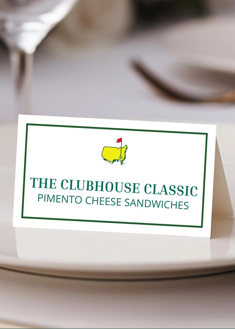 May include: A white card with a green border and text that reads "THE CLUBHOUSE CLASSIC PIMENTO CHEESE SANDWICHES." The card features a yellow outline of a golf course with a red flag. The card is on a white plate.