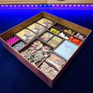 Könnte beinhalten: Ein hölzerner Spiel-Organizer mit mehreren Fächern, die mit Spielsteinen, Karten und Spielmarken gefüllt sind. Der Organizer enthält rosa, graue, orangefarbene und gelbe Spielsteine sowie Karten und Spielmarken. Der Organizer ist für die Aufbewahrung von Brettspielkomponenten konzipiert.