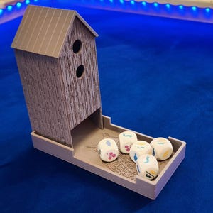 Op de afbeelding: Een lichtbruine houten dobbelsteentoren in de vorm van een klein huis, met twee ronde openingen en een dienblad. Zes witte dobbelstenen met bloemen- en geometrische patronen liggen in de lade. De toren en de lade staan op een blauw oppervlak.
