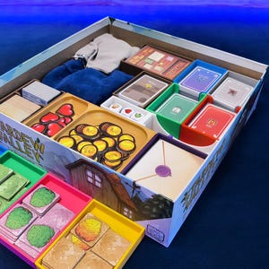 Pode incluir: A caixa do jogo de tabuleiro Stardew Valley está aberta, revelando compartimentos organizados cheios de componentes. Inclui fichas de coração vermelhas, moedas de ouro, vários baralhos de cartas, pequenos dados de madeira e tabuleiros de plástico coloridos com peças quadradas ilustradas com árvores, arbustos e terrenos.