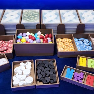 Op de afbeelding: Een verzameling bordspelcomponenten, waaronder kaarten, fiches en houten stukken, georganiseerd in bruine bakjes. De componenten zijn in verschillende kleuren, zoals roze, groen, rood, geel en blauw. De kaarten hebben een witte achtergrond met een blauw-wit ontwerp.