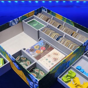 May include: A board game organizer with multiple compartments and trays. The main box is decorated with a colorful floral and leaf pattern. The organizer includes card slots, token holders, and separate trays for game pieces. Various game components are visible.