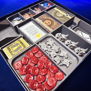 May include: A grey plastic board game organiser insert containing various game components. The tray features compartments filled with red circular tokens, silver miniature starships, white cards with text, and small stacks of game cards with gold and brown geometric patterns.