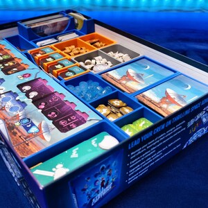 May include: A blue board game box with compartments containing game pieces, cards, and tokens. The box has colourful artwork and text, including the phrase "Lead Your Crew on Thrilling Space Adventures."