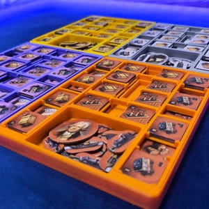 Può includere: Un set di quattro organizer per giochi da tavolo in plastica di colore giallo, grigio, viola e arancione. Ogni vassoio presenta scomparti multipli riempiti con vari gettoni di gioco in cartone quadrati e circolari, alcuni dei quali con ritratti.