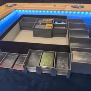 May include: A grey board game organiser with multiple compartments and trays. The organiser is designed to hold game pieces, cards, and other components. The trays are arranged in a tiered configuration, with various sizes and shapes to accommodate different game elements.