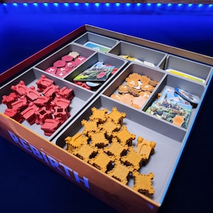 May include: A board game box labelled REBIRTH containing grey plastic storage compartments. The compartments hold red and orange castle-shaped game pieces, small red tokens, tan circular tokens, and rectangular game cards with colourful illustrations.