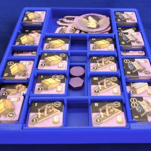 May include: A blue plastic game organizer with multiple compartments. The organizer holds game cards with illustrations of wooden crates and machinery, along with purple tokens and circular game pieces. The game pieces are arranged in a grid-like pattern.