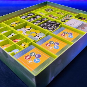 Puede incluir: Un organizador de juegos de mesa con interior verde lima y exterior verde claro. El organizador está dividido en múltiples compartimentos llenos de piezas de juego, cartas y fichas. Las cartas tienen diseños circulares coloridos.
