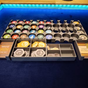 May include: A gray board game organizer with multiple compartments and trays. The organizer holds various game pieces, including colorful tokens and cards. The text "WAR ZON CHC SC" is visible on one tray, and "ROYAL DECREE" on another.