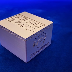 Puede incluir: Una caja cuadrada de color claro con las palabras "SEA SALT & PAPER" en relieve en la tapa. El lateral de la caja presenta un personaje de dibujos animados de setas y el texto "ShroomForge". La caja está sobre un fondo azul.