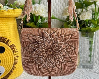 May include: A beaded evening bag in a warm rose gold hue, featuring a large floral design on the front. The bag has a matching beaded strap and tassel accents. The bag is displayed on a stand with a white background and flowers.
