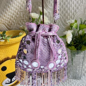 May include: A lavender drawstring bag with a beaded handle and fringe. The bag is decorated with small, round mirrors and decorative beads. The bag is displayed on a metal stand.