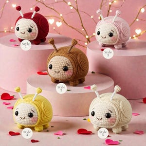 May include: Five crocheted bug-like toys in various colours: red, brown, pink, yellow, and cream. Each has a smiling face, small legs, and antennae. The toys are displayed on pink platforms with heart-shaped confetti.