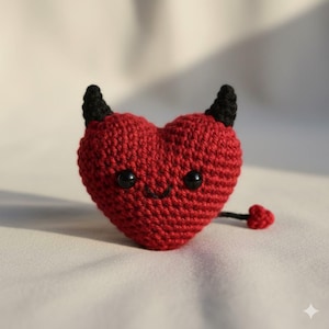 Op de afbeelding: Een handgemaakt rood gehaakt hart met zwarte hoorns, een kleine staart en een lachend gezicht. Het hartvormige pluche heeft zwarte knoogogen en een gestikte mond. Het duivel-thema item is een unieke, grillige creatie.