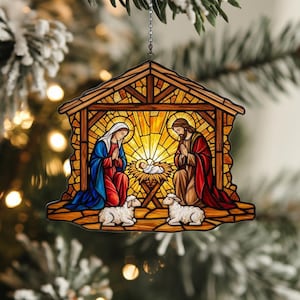 May include: A stained-glass style Christmas ornament depicting the Nativity scene. The ornament features Mary and Joseph kneeling beside baby Jesus in a manger, with two sheep. The ornament is hanging from a silver chain.