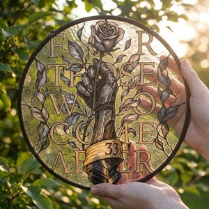 May include: Circular stained glass artwork in a black frame. The design shows a mechanical arm holding a rose, with the words "For Those Who Come After" and the number "33" etched into the glass. The artwork is held against a backdrop of green foliage.