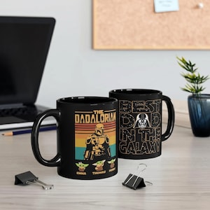 K&ouml;nnte beinhalten: Schwarze Keramik-Tasse mit "The Dadalorian"-Design, mit einer mandalorianischen Figur und Grogu-Charakteren. Die andere Seite zeigt "Best Dad in the Galaxy" mit Darth Vader. Ein einzigartiges Geschenk f&uuml;r Star Wars-Fans.