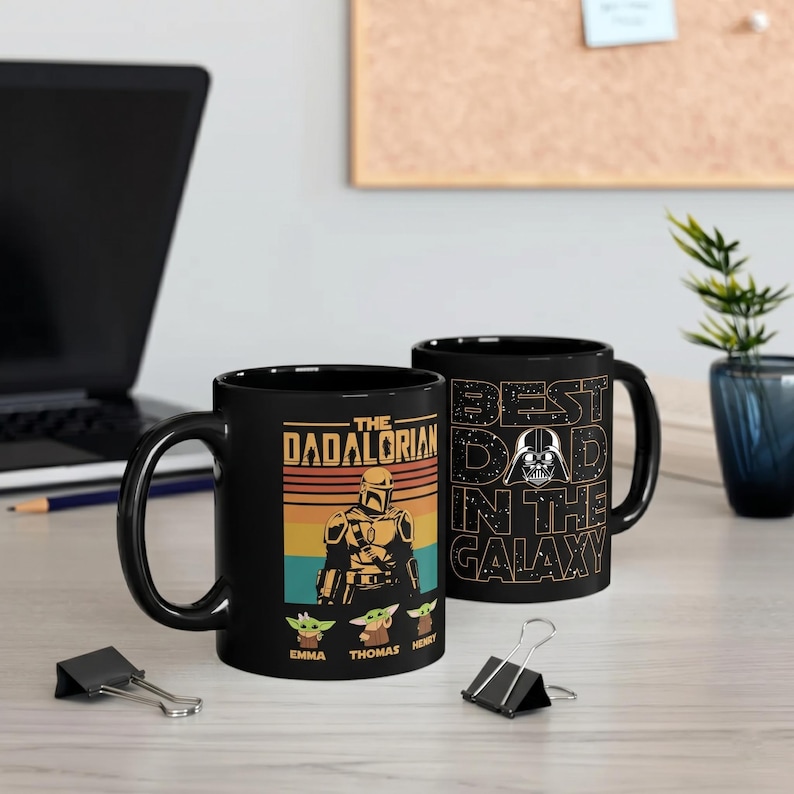K&ouml;nnte beinhalten: Schwarze Keramik-Tasse mit "The Dadalorian"-Design, mit einer mandalorianischen Figur und Grogu-Charakteren. Die andere Seite zeigt "Best Dad in the Galaxy" mit Darth Vader. Ein einzigartiges Geschenk f&uuml;r Star Wars-Fans.