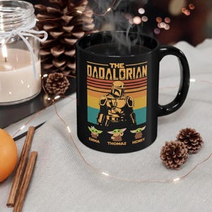K&ouml;nnte beinhalten: Schwarze Keramik-Tasse mit dem Design "The Dadalorian", das eine mandalorianische Figur und drei Grogu-Charaktere zeigt. Die Tasse tr&auml;gt die Namen "Emma", "Thomas" und "Henry" unter den Grogu-Figuren. Das Design ist vor einem Retro-Sonnenuntergangshintergrund.