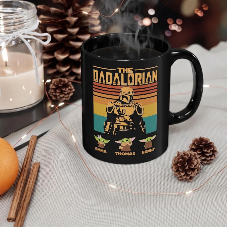 K&ouml;nnte beinhalten: Schwarze Keramik-Tasse mit dem Design "The Dadalorian", das eine mandalorianische Figur und drei Grogu-Charaktere zeigt. Die Tasse tr&auml;gt die Namen "Emma", "Thomas" und "Henry" unter den Grogu-Figuren. Das Design ist vor einem Retro-Sonnenuntergangshintergrund.