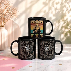 K&ouml;nnte beinhalten: Drei schwarze Keramikbecher mit Star Wars-Designs. Ein Becher zeigt "THE DADALORIAN" mit einer Charakterillustration und den Namen "EMMA, THOMAS, HENRY". Die anderen beiden Becher zeigen "BEST DAD IN THE GALAXY" mit einer Darth-Vader-Grafik.