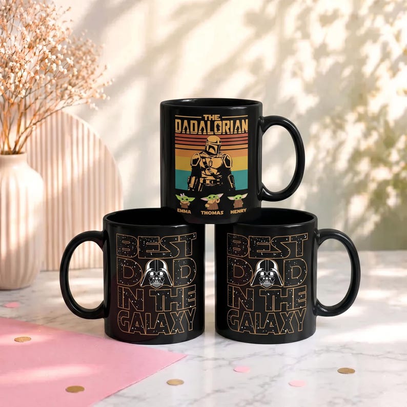 K&ouml;nnte beinhalten: Drei schwarze Keramikbecher mit Star Wars-Designs. Ein Becher zeigt "THE DADALORIAN" mit einer Charakterillustration und den Namen "EMMA, THOMAS, HENRY". Die anderen beiden Becher zeigen "BEST DAD IN THE GALAXY" mit einer Darth-Vader-Grafik.