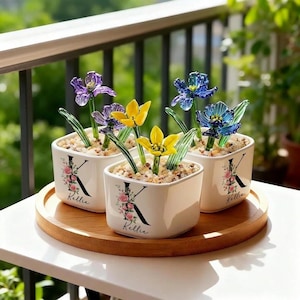 May include: Three square white ceramic planters with glass flower sculptures. The planters have a floral design with the letter "K" and the name "Kellie". The flowers are purple, yellow, and blue.