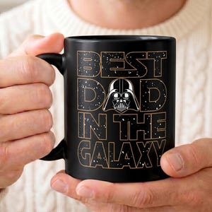 K&ouml;nnte beinhalten: Schwarze Keramik-Tasse mit dem Text "BEST DAD IN THE GALAXY" in einer verwitterten Schriftart, mit einer Darth Vader-Grafik. Die Tasse hat einen schwarzen Henkel und wird von einer Person gehalten.
