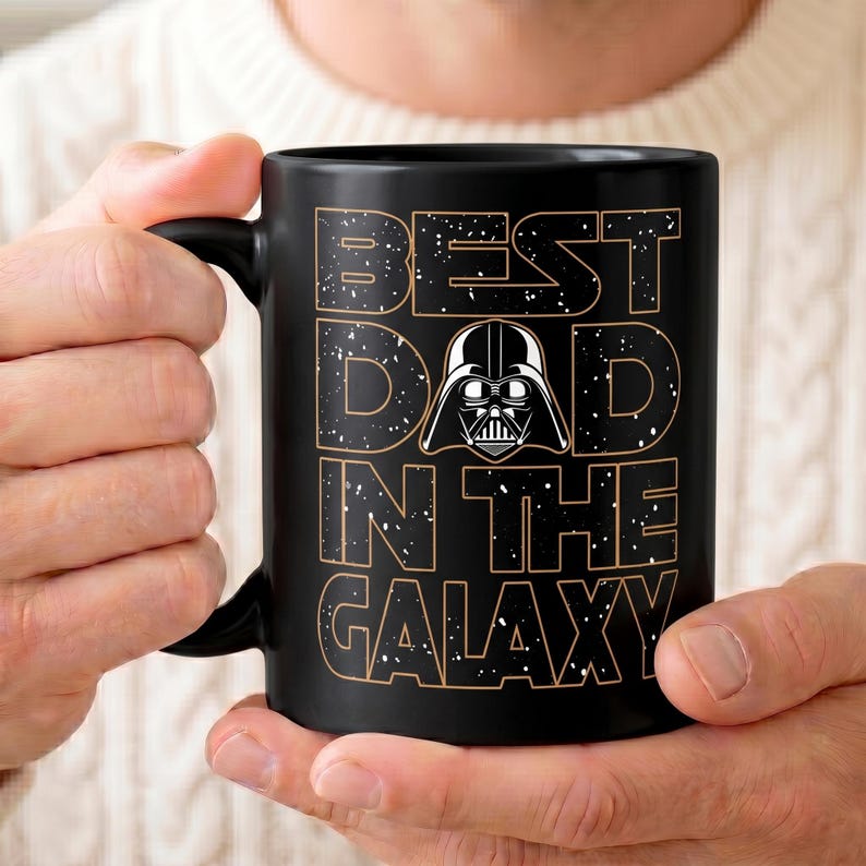 K&ouml;nnte beinhalten: Schwarze Keramik-Tasse mit dem Text "BEST DAD IN THE GALAXY" in einer verwitterten Schriftart, mit einer Darth Vader-Grafik. Die Tasse hat einen schwarzen Henkel und wird von einer Person gehalten.