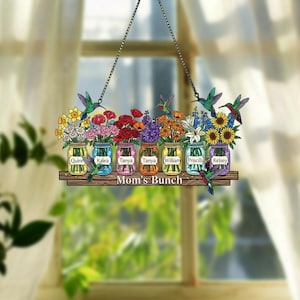 May include: A decorative hanging sign featuring a wooden shelf with six mason jars filled with colorful flowers. Each jar has a name on it. Hummingbirds are flying around the flowers. The sign reads "Mom's Bunch".