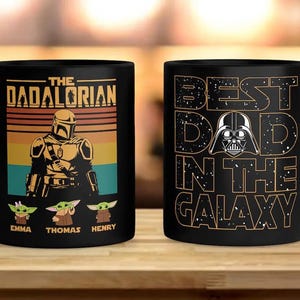 K&ouml;nnte beinhalten: Zwei schwarze Keramikbecher. Ein Becher zeigt den Text "THE DADALORIAN" mit einer mandalorianischen Figur und drei Grogu-Figuren. Der andere Becher sagt "BEST DAD IN THE GALAXY" mit einer Darth Vader-Grafik.