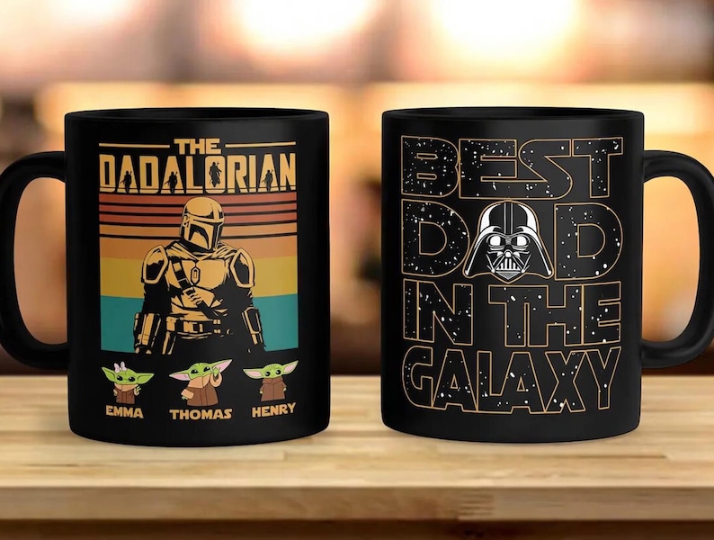 K&ouml;nnte beinhalten: Zwei schwarze Keramikbecher. Ein Becher zeigt den Text "THE DADALORIAN" mit einer mandalorianischen Figur und drei Grogu-Figuren. Der andere Becher sagt "BEST DAD IN THE GALAXY" mit einer Darth Vader-Grafik.