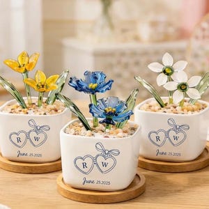 May include: Three small white ceramic pots with glass flowers in yellow, blue, and white. Each pot has a heart design with the initials "R" and "W" and the date "June 25.2025". The pots sit on wooden coasters.