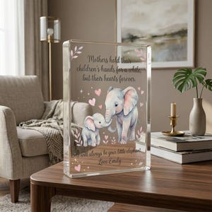 May include: A clear acrylic block features a watercolor illustration of two elephants, one adult and one baby, with pink ears. The block has text that reads, "Mothers hold their children's hands for a while, but their hearts forever."