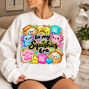 Puede incluir: Una sudadera blanca de cuello redondo con un estampado gráfico colorido con el texto In my Squishies Era. El diseño incluye ajolotes, dumplings en vaporeras de bambú, una barra de mantequilla, huellas y estrellas en colores brillantes.