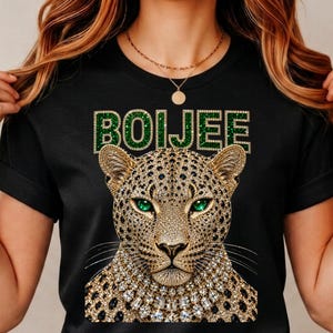 May include: A black short-sleeved t-shirt featuring a graphic of a leopard with emerald green eyes and a jewelled collar. Above the leopard, the word BOIJEE is printed in a green, glittery, block-letter font with a gold-toned outline.