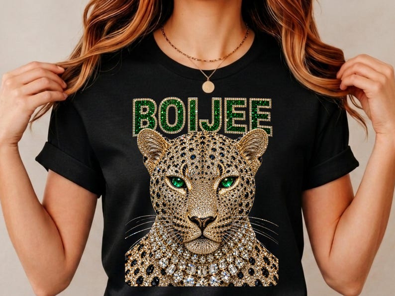 May include: A black short-sleeved t-shirt featuring a graphic of a leopard with emerald green eyes and a jewelled collar. Above the leopard, the word BOIJEE is printed in a green, glittery, block-letter font with a gold-toned outline.