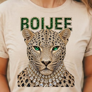 May include: A beige t-shirt featuring a graphic of a leopard head with emerald green eyes and a jeweled collar. The word BOIJEE is printed above the leopard in a green, glittery, block-letter font. The leopard design is covered in gold and black spots.