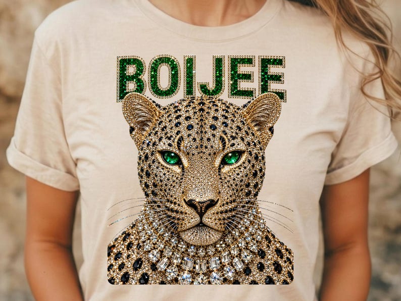 May include: A beige t-shirt featuring a graphic of a leopard head with emerald green eyes and a jewelled collar. The word BOIJEE is printed above the leopard in a green, glittery, block-letter font. The leopard design is covered in gold and black spots.