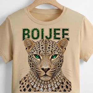 May include: A beige short-sleeved t-shirt featuring a graphic of a leopard head with green eyes and a crystal-style necklace. Above the leopard, the word BOIJEE is printed in a green, glittery, textured font. The design is centred on the chest.