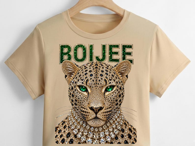 May include: A beige short-sleeved t-shirt featuring a graphic of a leopard head with green eyes and a crystal-style necklace. Above the leopard, the word BOIJEE is printed in a green, glittery, textured font. The design is centred on the chest.