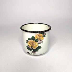 May include: A white enamel mug with a black rim and a floral design. The design features yellow roses with green leaves and small white flowers. The mug has a slightly distressed appearance.