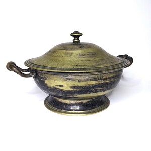 May include: A vintage metal serving bowl with a lid. The bowl has a pedestal base and two handles. The exterior is a mix of gold and black, with a decorative knob on the lid. The bowl is likely used for serving soups or stews.