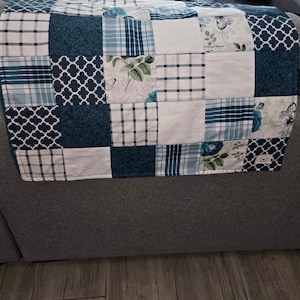 May include: A patchwork quilt with a variety of blue and white patterns. The quilt features squares of solid colors, plaid, floral, and geometric designs. Butterflies and floral motifs are also present. The quilt is draped over a gray sofa.