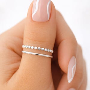 Puede incluir: Dos anillos de plata se llevan en un dedo, uno con pequeños acentos circulares y el otro una banda simple. Las uñas están pintadas de rosa claro con líneas onduladas blancas. El fondo es blanco suave.